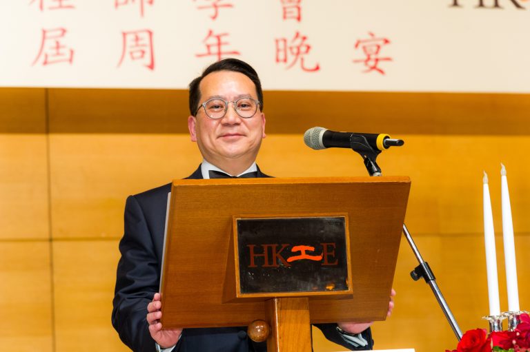 Man in suit and glasses speaking at podium with HKIE logo during anniversary banquet.